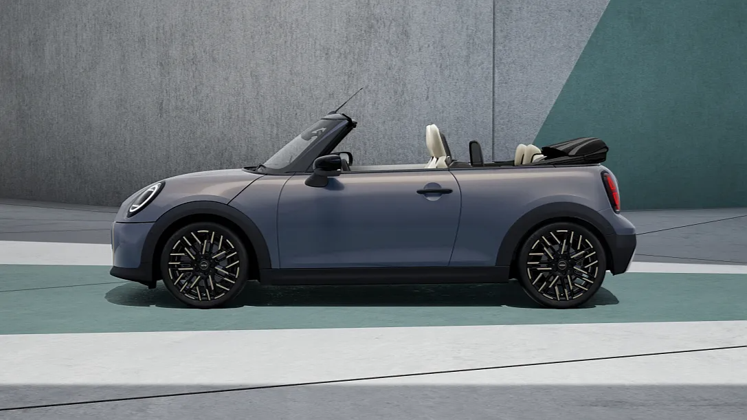 2025 MINI Convertible side profile in grey with the roof down, showcasing its compact design and black alloy wheels in an outdoor modern setting.