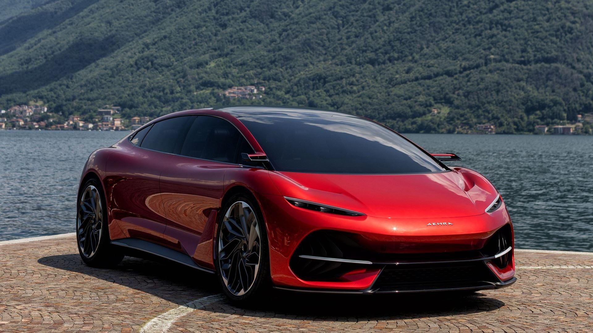 A red AEHRA electric sedan concept car parked beside a lake, featuring a sleek aerodynamic design, large alloy wheels, and a panoramic glass roof with mountains in the background.