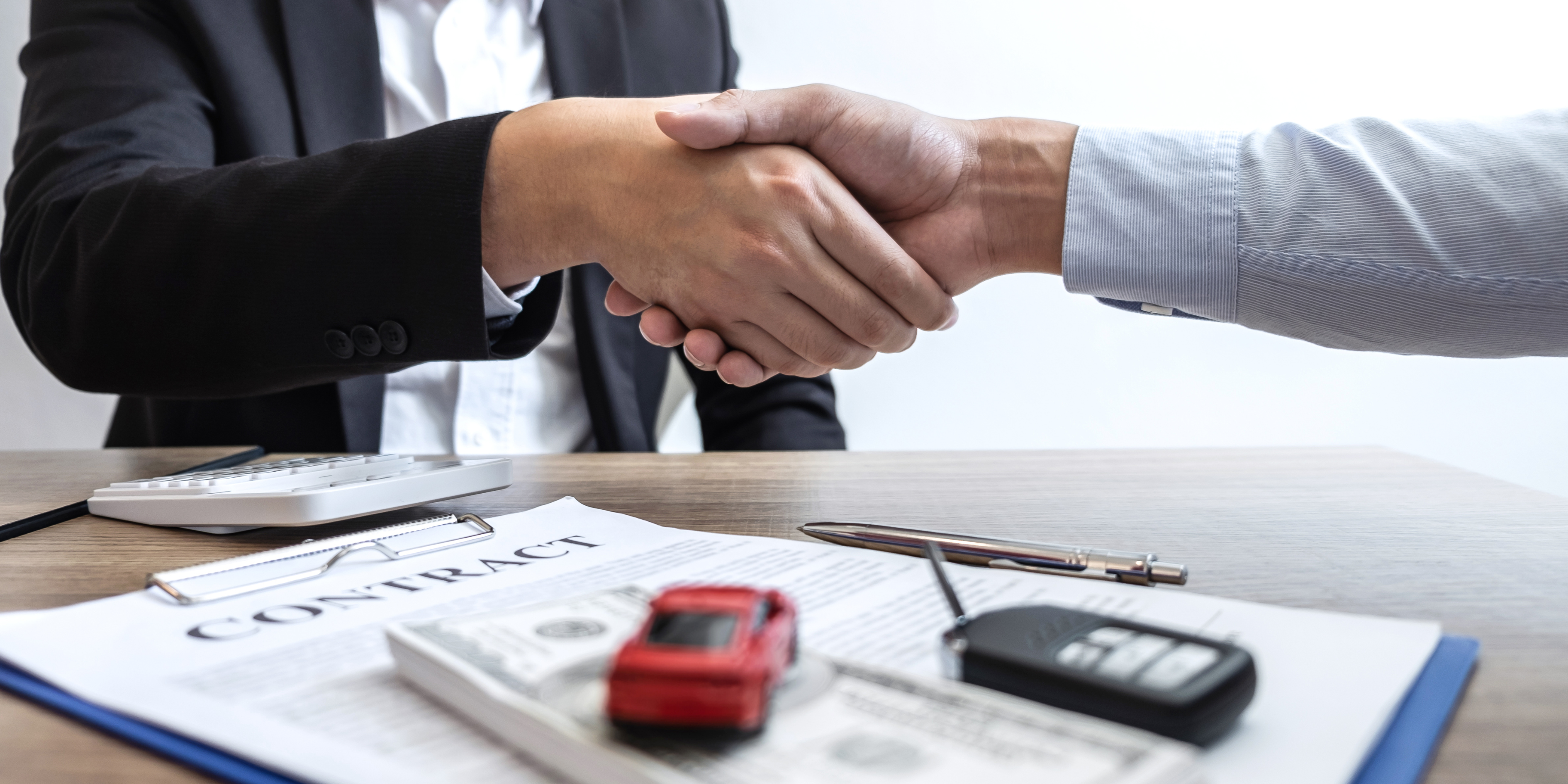 Car buyer and dealer shaking hands over a signed vehicle purchase contract with car keys, calculator, and model car on the desk.