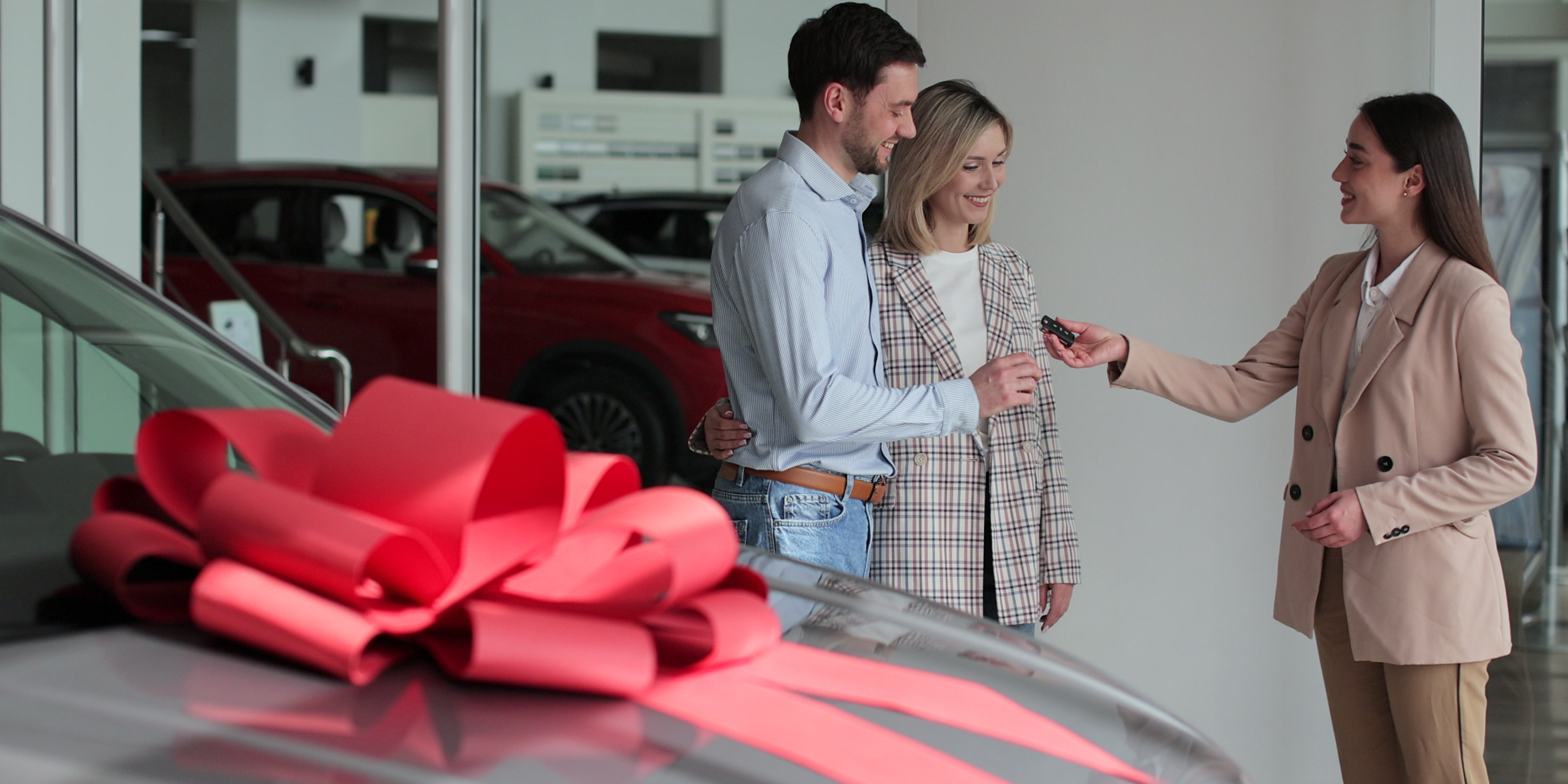 Car salesperson handing keys to a couple inside a showroom with a new car decorated with a red bow.