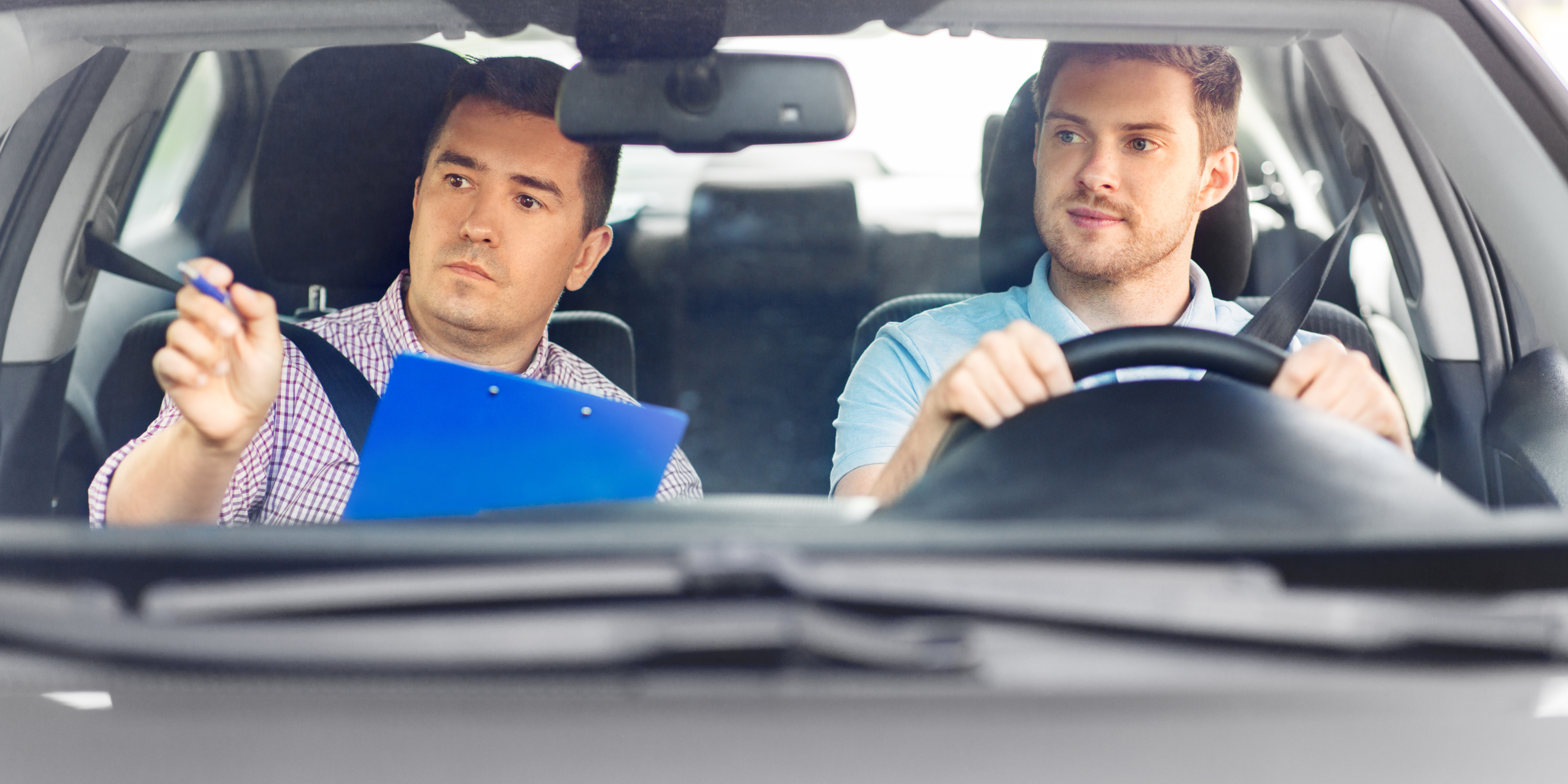 Man test-driving a car while an instructor evaluates with a clipboard.